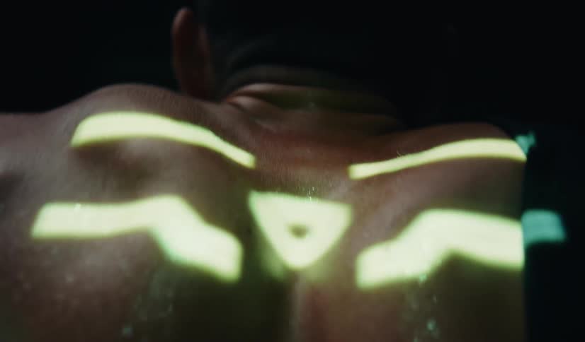Abstract neon green symbols and patterns are projected onto the muscular back of a person in a dark studio