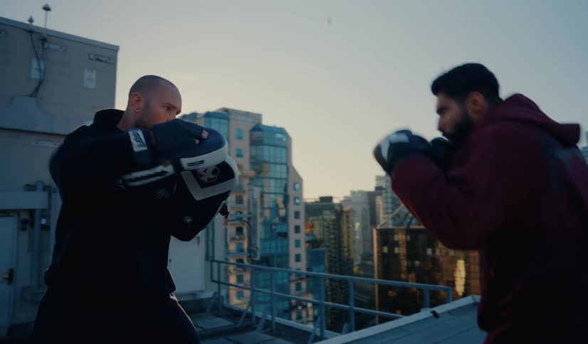 A boxer and his coach train with focus mitts on a city rooftop with a skyline view at sunset