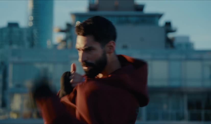 A determined male boxer in a red hoodie intensely shadowboxes on a city rooftop at dawn