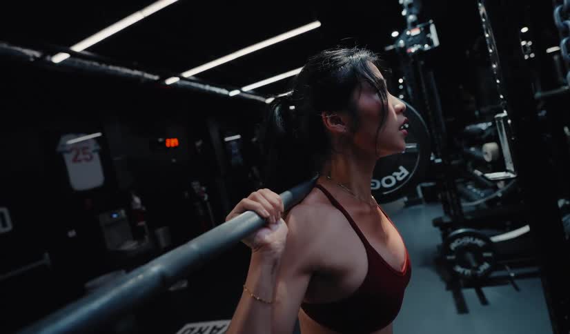 Slow-motion side profile of a fit athletic woman doing squats with a barbell in a dark and moody gym