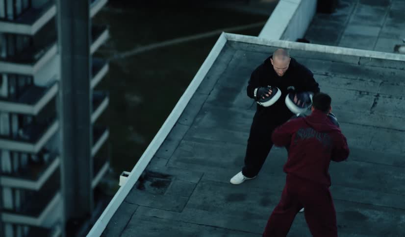 An overhead view shows a boxer and his coach training with focus mitts on a city rooftop
