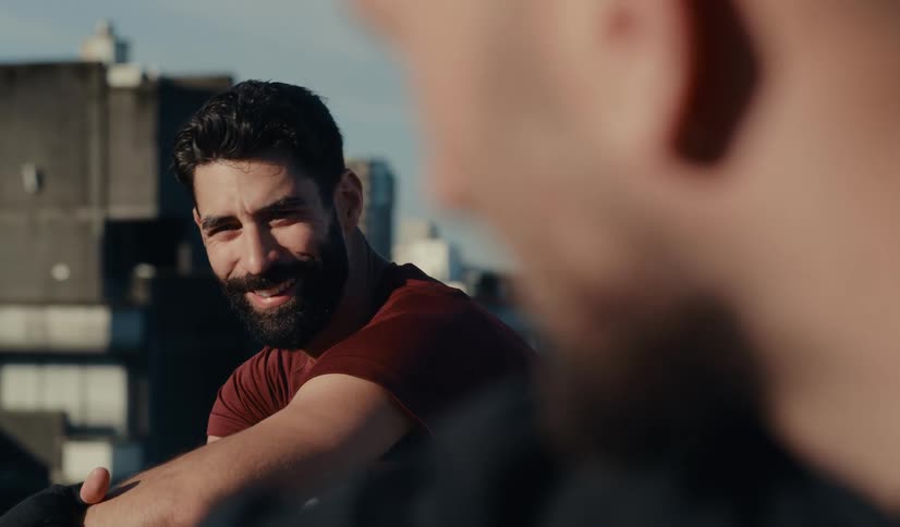 Over-the-shoulder shot of a bearded man smiling while talking to his friend on a city rooftop on a sunny day