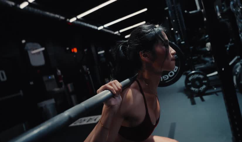 A determined woman performs a slow-motion barbell squat in a dark, modern gym