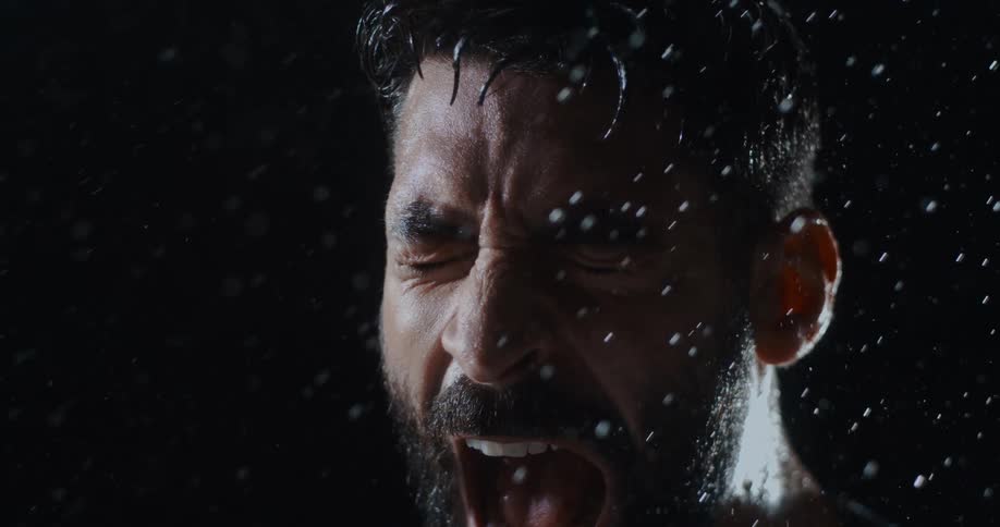 Cinematic slow motion close-up of a bearded man screaming in anguish in the pouring rain against a black background
