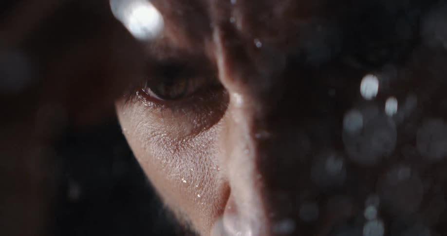 A dramatic slow-motion close-up reveals a man's intense gaze as water droplets fall on his face in the dark