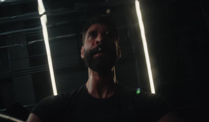 Low-angle slow motion shot of a man's face as he spits and splashes water while intensely working out in a dark, modern gym