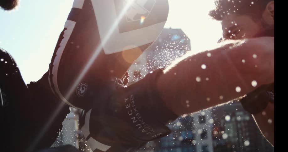 In a dramatic slow-motion close-up, a boxer's gloved fist strikes a training mitt, sending a spray of sweat into the sunlit city air