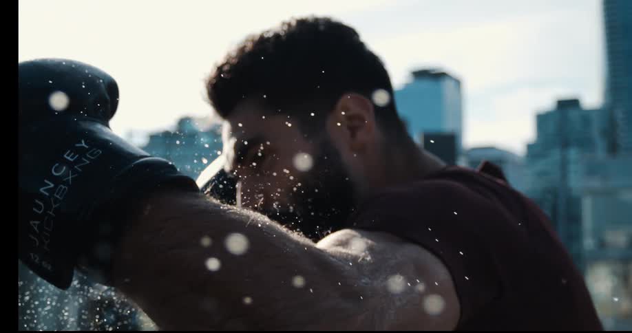 A determined boxer punches a focus mitt in slow motion, sending beads of sweat flying through the air with the city skyline in the background