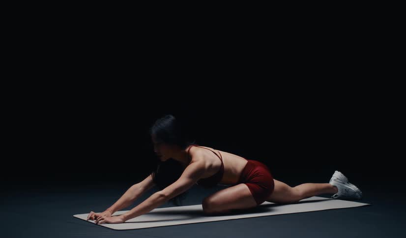 A fit Asian woman in a dark studio slowly performs a deep pigeon pose stretch on a yoga mat