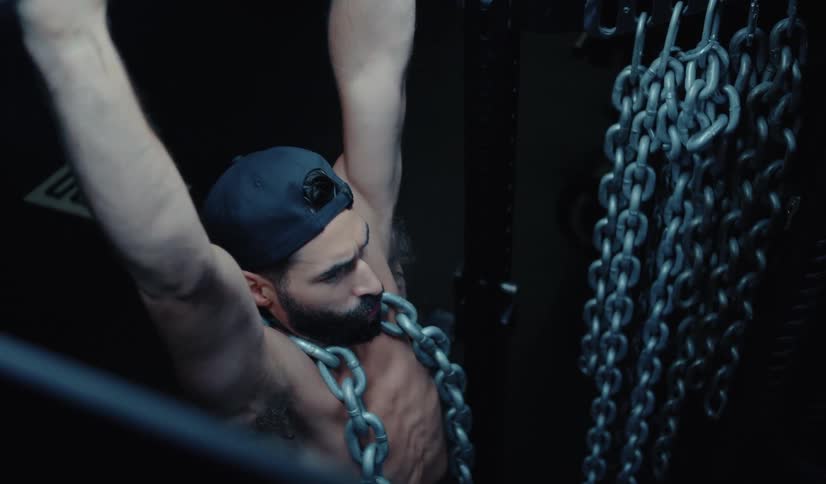 A muscular, bearded man wearing a backwards cap performs an intense weighted pull-up with a heavy metal chain in a dark gym