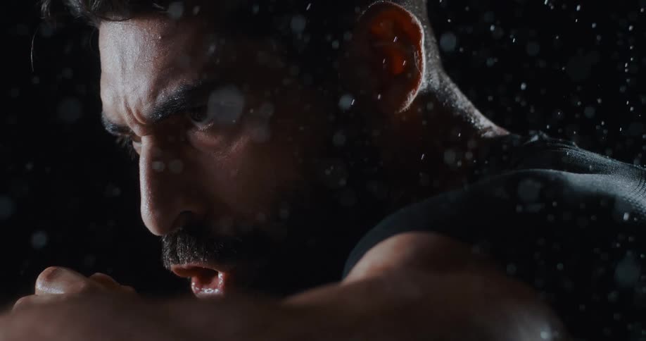 An extremely determined and focused man tenses his muscles and breathes hard while being drenched in water in super slow motion