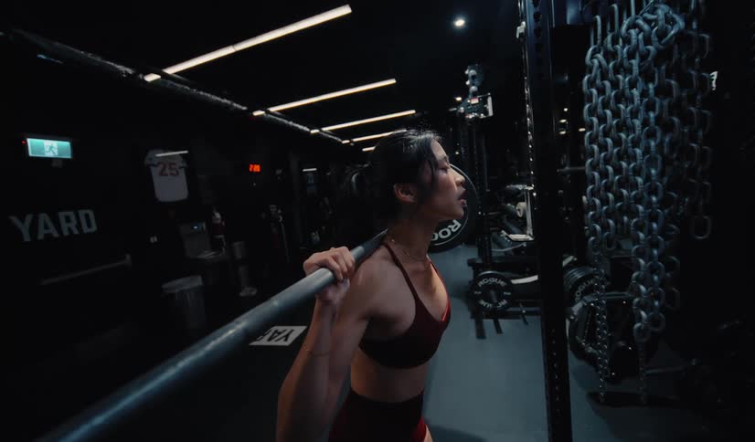 A focused and determined woman performs a slow-motion barbell squat in a dark, atmospheric gym