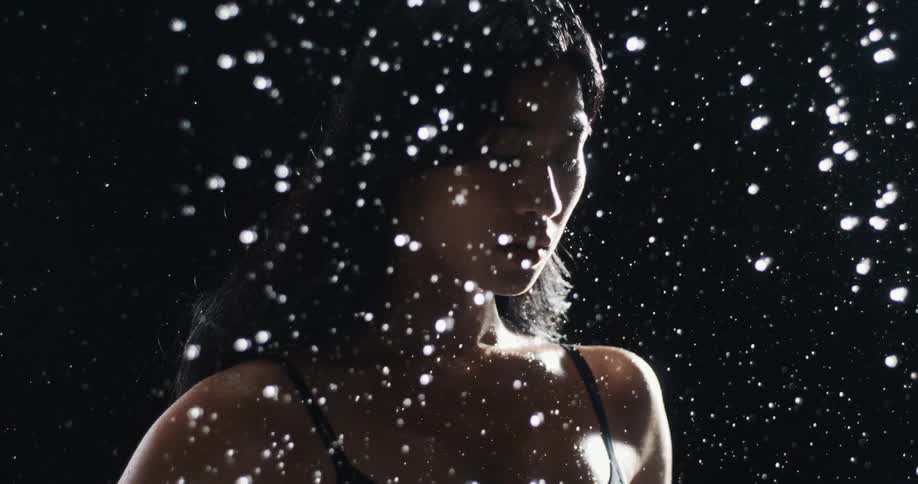 Cinemagraph of a serene woman standing still behind a curtain of backlit water droplets falling against a dark background