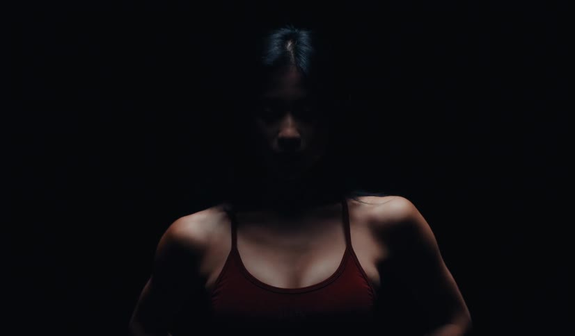 A fit Asian woman with her eyes closed in a red sports bra gracefully stretches her neck in slow motion against a dramatic black background