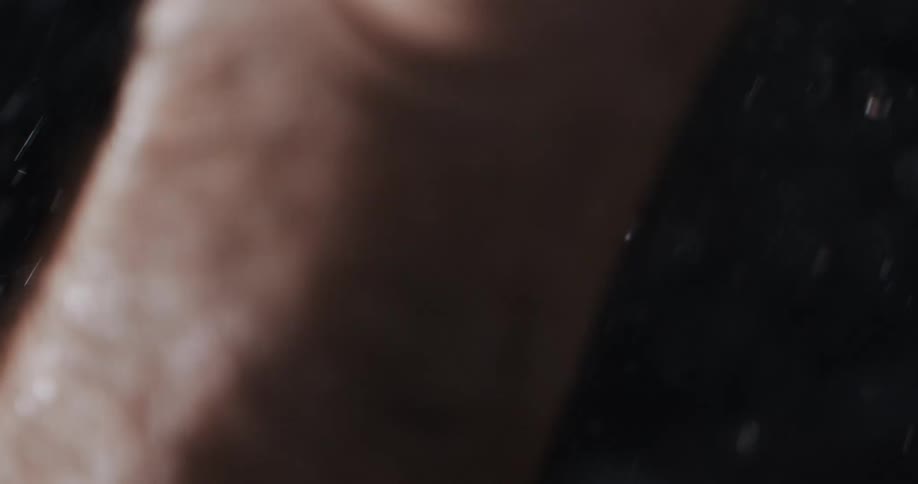 An intimate macro shot in super slow motion captures water droplets falling onto a person's hand and body against a dark background
