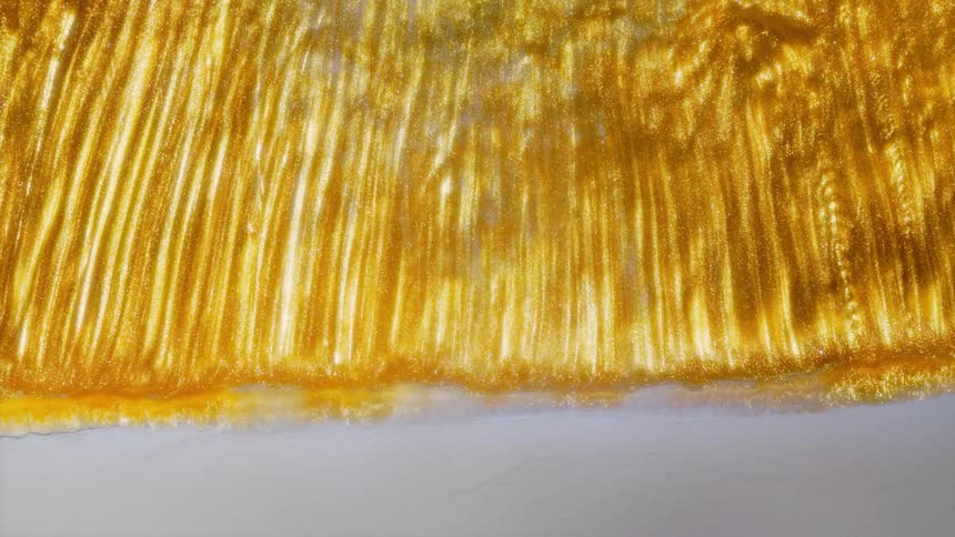 A close-up view of thick, shimmering golden liquid as it slowly flows and drips down a pristine white surface