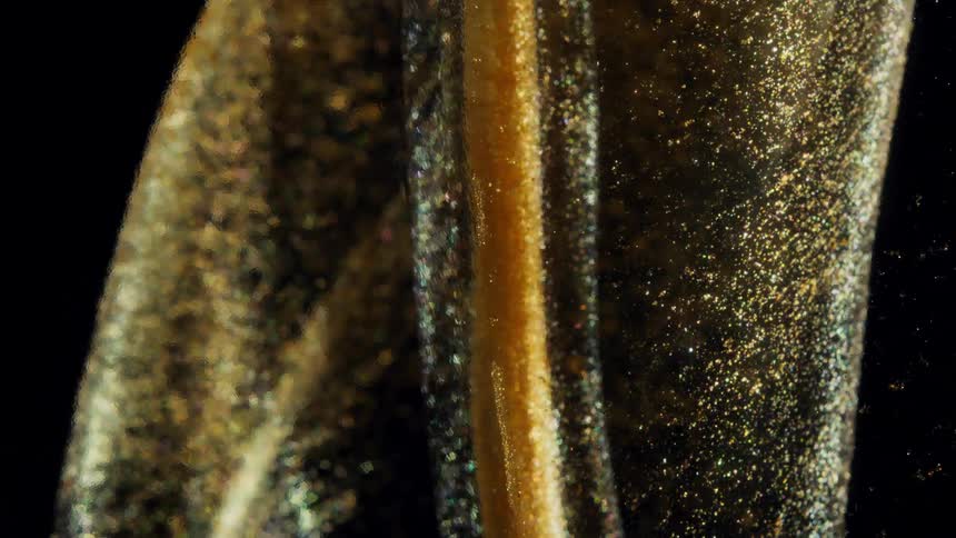 A macro shot of a viscous golden fluid with shimmering glitter slowly flowing against a black backdrop
