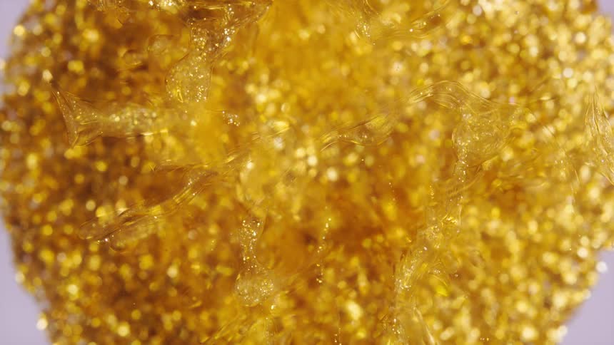A shimmering macro view shows golden glitter suspended and swirling within a clear, viscous liquid