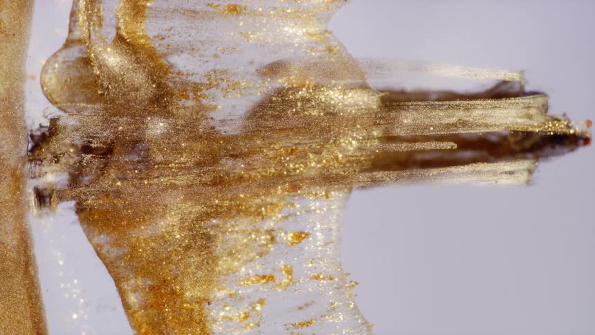 A vertical macro shot of golden particles slowly flowing and expanding in a clear viscous liquid against a white background