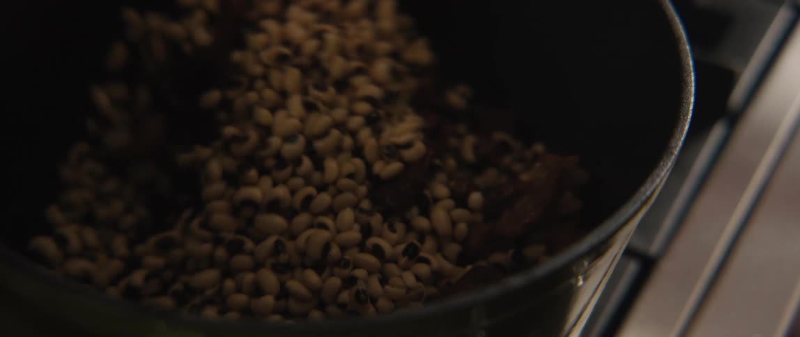 A close-up slow-motion shot of a person using a wooden spoon to stir black-eyed peas and meat in a pot
