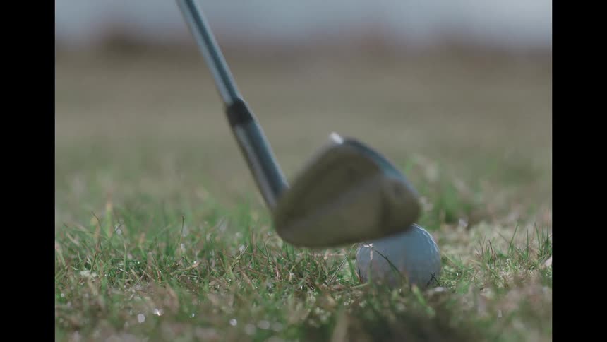 A slow-motion close-up captures a golf iron striking a ball on a dewy fairway, sending turf flying