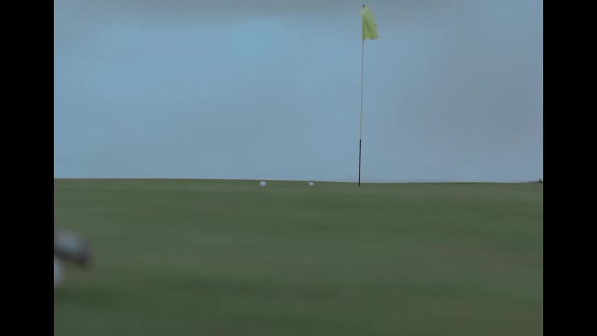 A slow-motion view from behind a golf club as it putts a ball across a green toward the hole and flag on an overcast day