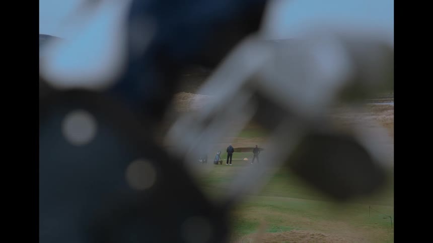 A distant view of men playing golf on a green course, framed by a blurry golf bag in the foreground
