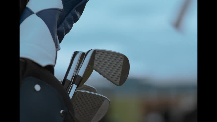 A gloved hand pulls a golf club from a bag, with a rack focus to the golf carts in the background