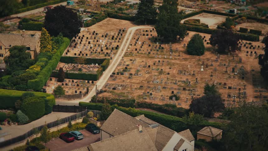 A solitary figure in black walks slowly through a quiet English village cemetery in a somber high-angle shot