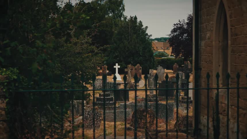 A slow, moody pan across a historic English church graveyard with weathered tombstones behind an old iron fence
