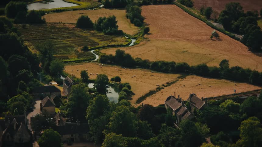 A time-lapse aerial view of an idyllic English countryside village with sheep grazing in fields next to a winding river as shadows move across the landscape