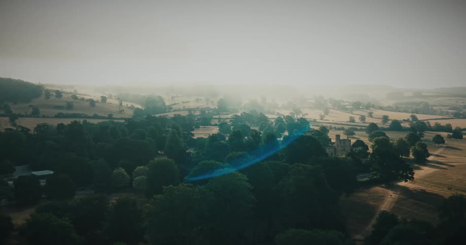 A dreamy aerial view shows a historic castle nestled amongst the trees in a vast, misty countryside landscape as the sun creates a beautiful lens flare