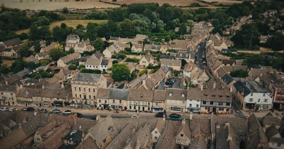 An ascending aerial shot reveals a picturesque and historic English countryside town with stone buildings and a bustling main street
