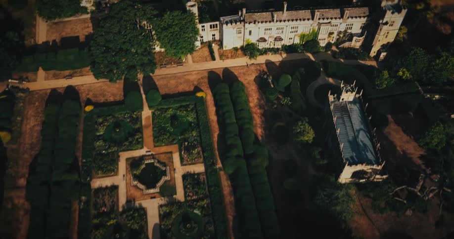 An aerial drone flies backwards over a grand historic castle with a central courtyard, revealing its expansive formal gardens and an adjacent chapel on a sunny day