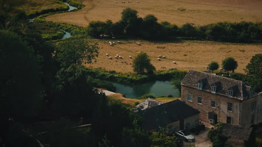 An idyllic high-angle view of a flock of sheep grazing in a large field next to a winding river and an old stone farmhouse in the countryside