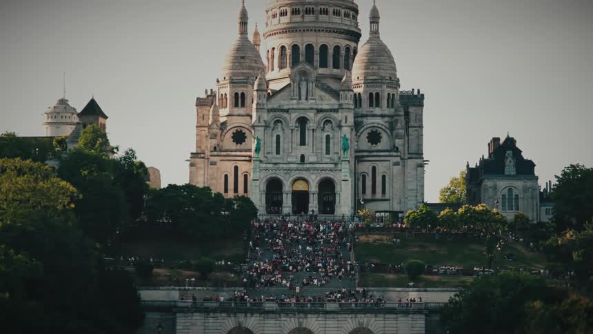 A slow zoom shot captures a large crowd of people gathered on the steps of the magnificent Sacré-Cœur Basilica in Montmartre, Paris