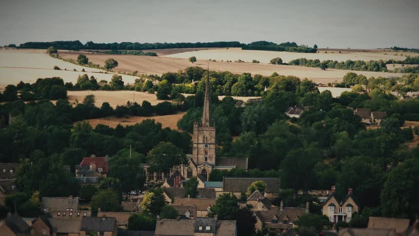A slow zoom out from a historic church steeple in a quaint English village surrounded by lush trees and rolling countryside