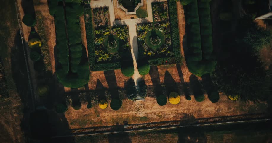 A dramatic aerial top-down view reveals an ornate and symmetrical formal garden with manicured hedges and fountains