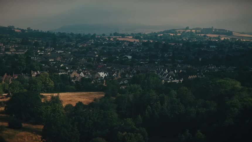 A slow aerial zoom shot reveals a picturesque countryside town nestled among rolling green hills and forests on a moody, overcast day