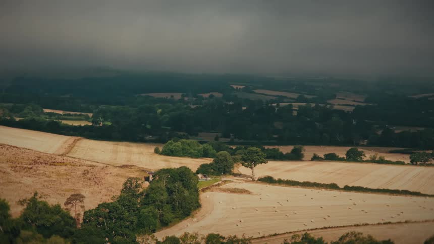 A dramatic aerial view of a rolling rural landscape with fields and forests under dark, moody storm clouds