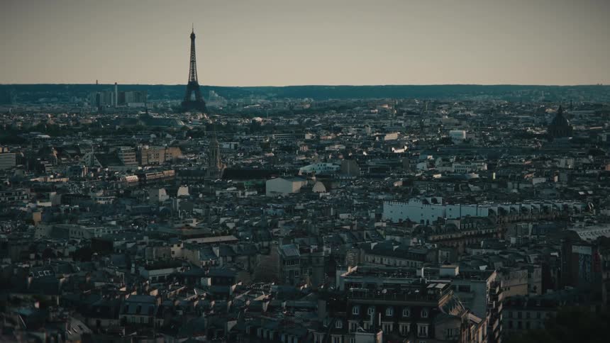 Slow aerial push-in shot over the rooftops of Paris, France, with the Eiffel Tower standing tall in the hazy distance at dusk
