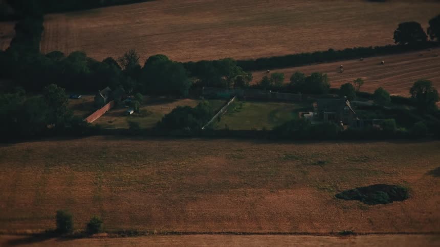 An aerial view slowly zooms towards a secluded farmhouse surrounded by vast, sun-drenched fields in the countryside