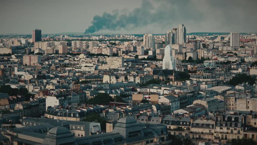 Slow zoom aerial shot of a dense cityscape with a large plume of black smoke rising in the distance
