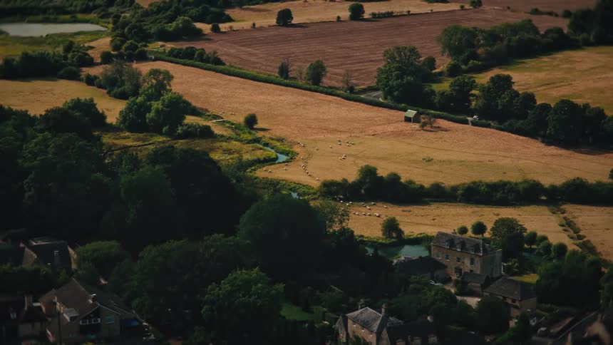 High angle view zooms out from a flock of sheep by a river to reveal a quaint English village nestled in the countryside