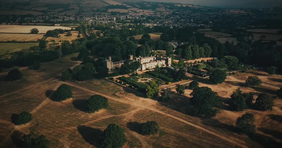 A majestic aerial pullback shot reveals a historic castle and its beautiful formal gardens, with the surrounding rural landscape and a distant town coming into view