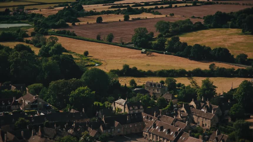 A slow aerial zoom captures a quaint European village with a historic church, nestled in a vast, rolling countryside with grazing sheep
