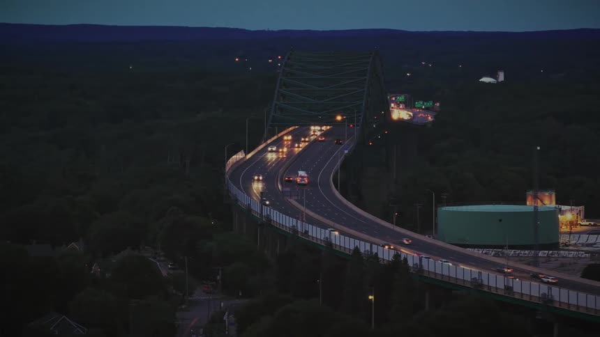 An aerial drone shot slowly reveals a curving highway bridge with steady traffic flow at dusk, set against a dark, forested landscape