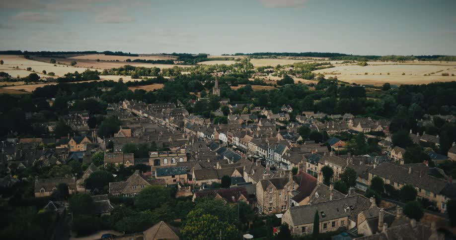 A cinematic aerial view slowly pulls back from a historic English countryside village nestled among rolling hills and fields