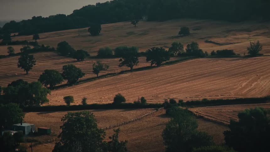 An aerial view slowly zooms in on vast, golden harvested farm fields nestled among the rolling hills of the countryside at sunset