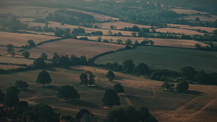 High angle slow zoom over a beautiful English countryside landscape with rolling hills, patchwork fields, and ancient ruins in the warm morning sun